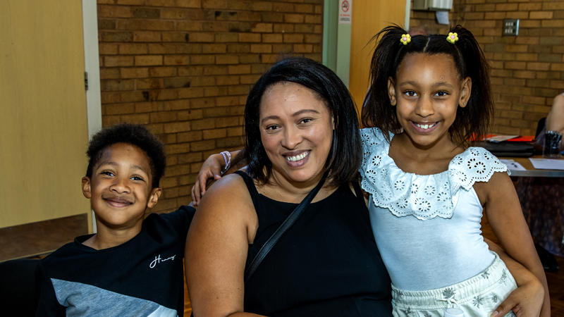 A woman is sat at a table. Her two children, a young boy to her left and young girl to her right, are stood next to her.