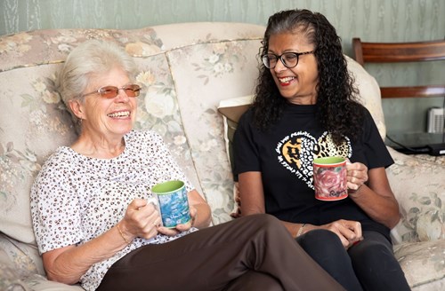 Two women are sat on a sofa. They are both holding mugs and are laughing.