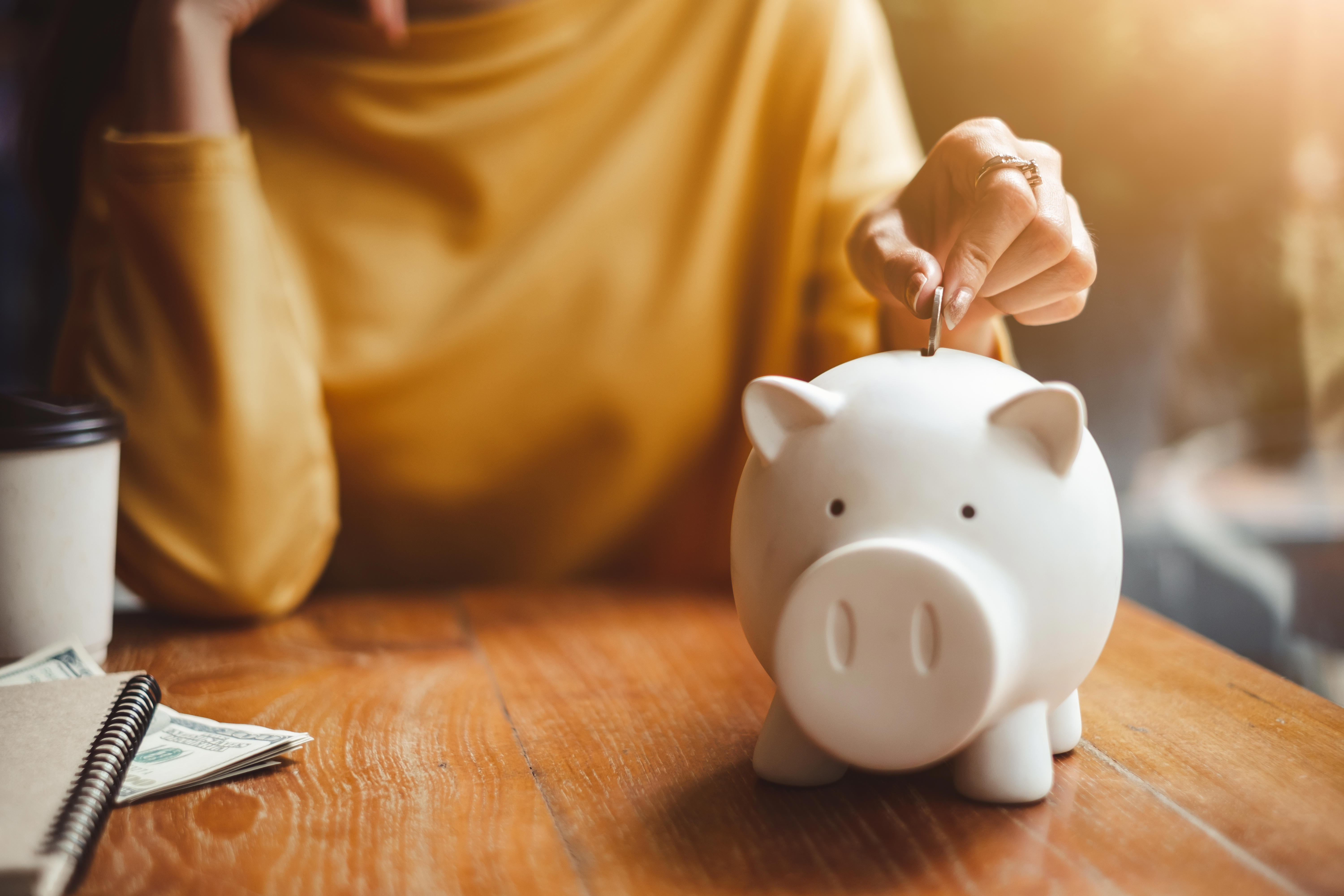 A white piggy bank, shaped like a pig, is having a coin put into it by a hand.