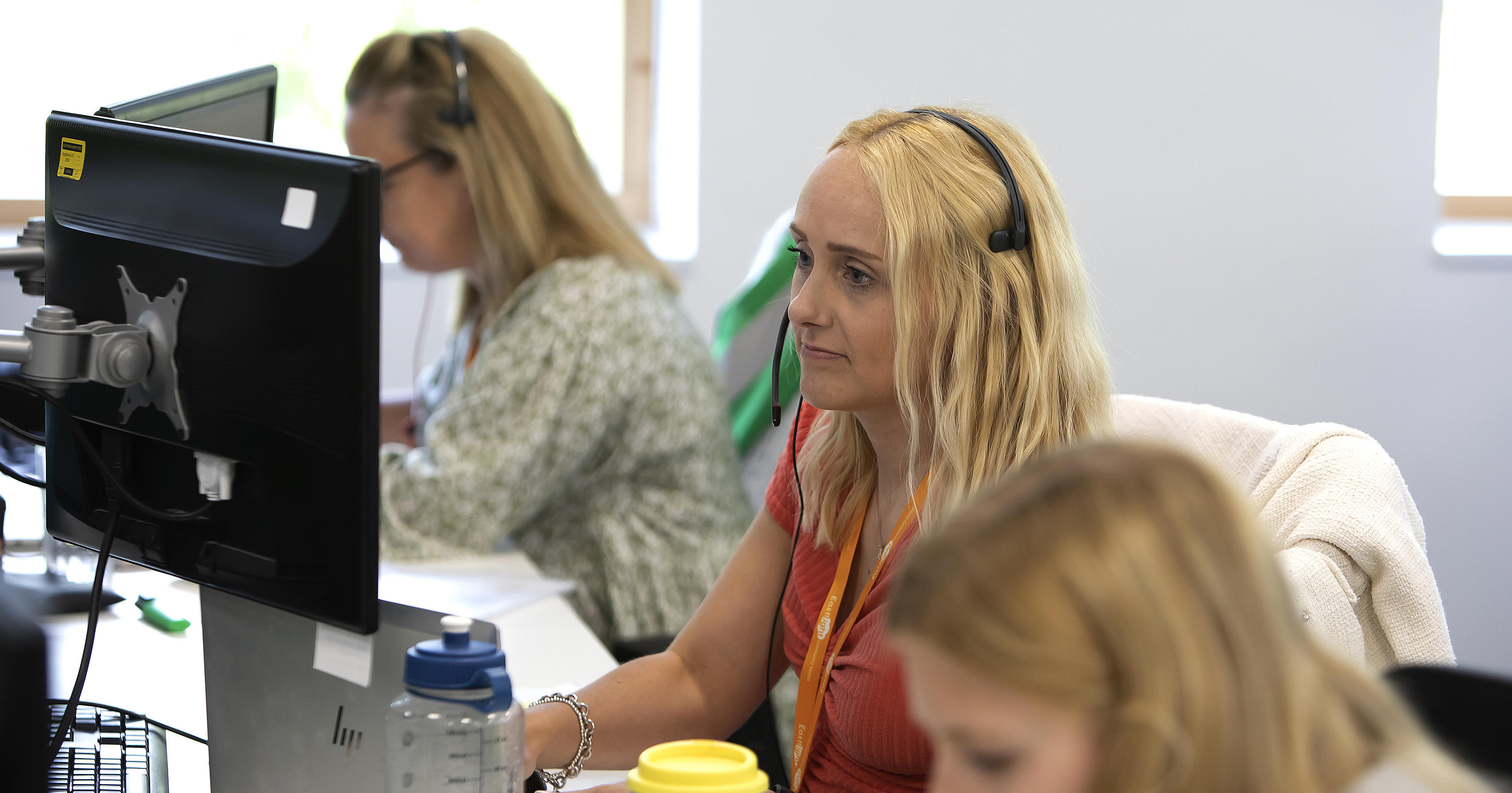 A woman wearing a headset is looking at a laptop screen. There's another woman in the background doing the same.