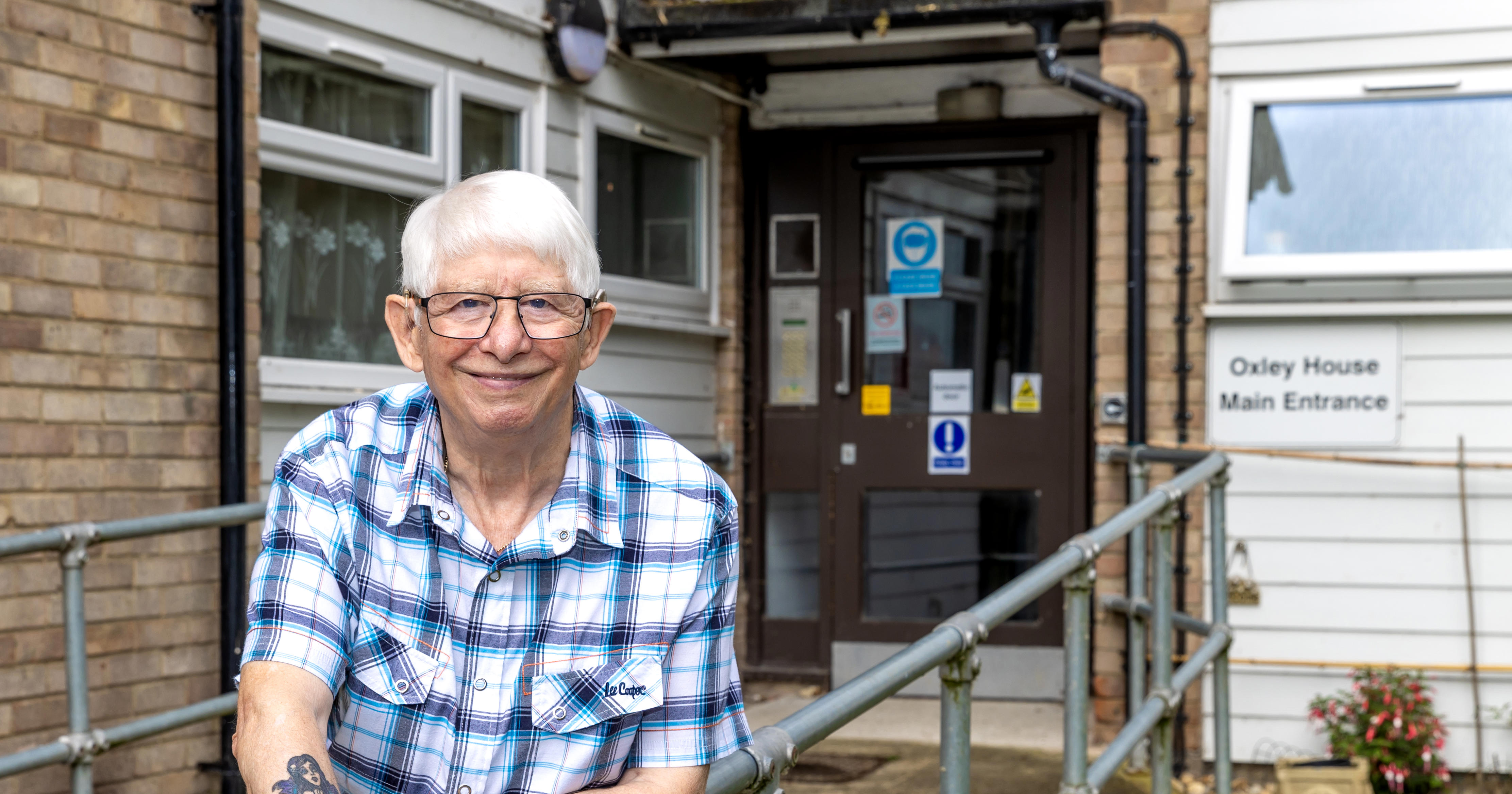 An older man with glasses is leaning on a metal railing which leads up a ramp to a block of flats.