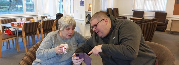 A man and a woman are looking at a phone. The man is pointing to the screen while the woman is holding a mug.