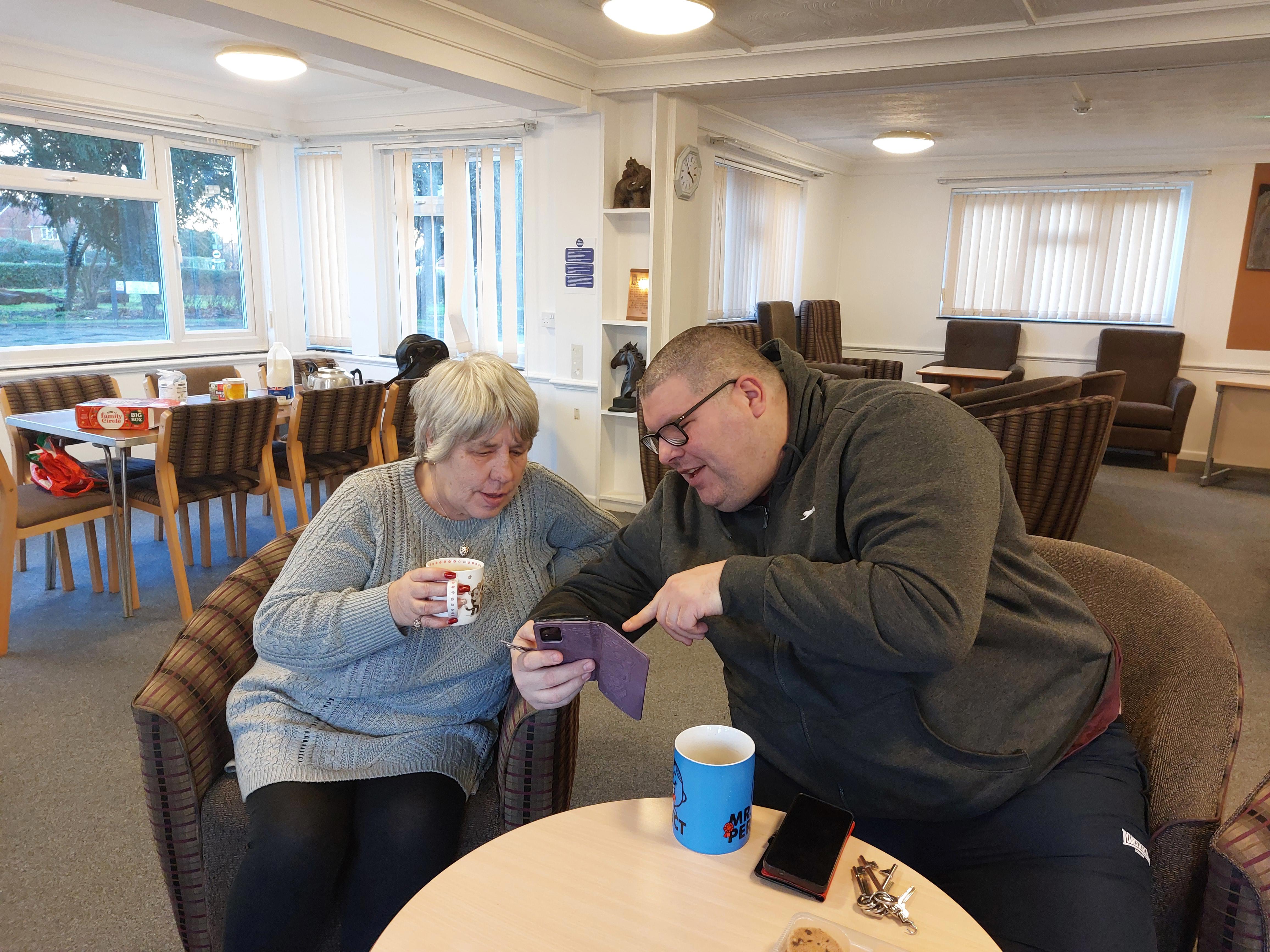 A man and a woman are looking at a phone. The man is pointing to the screen while the woman is holding a mug.