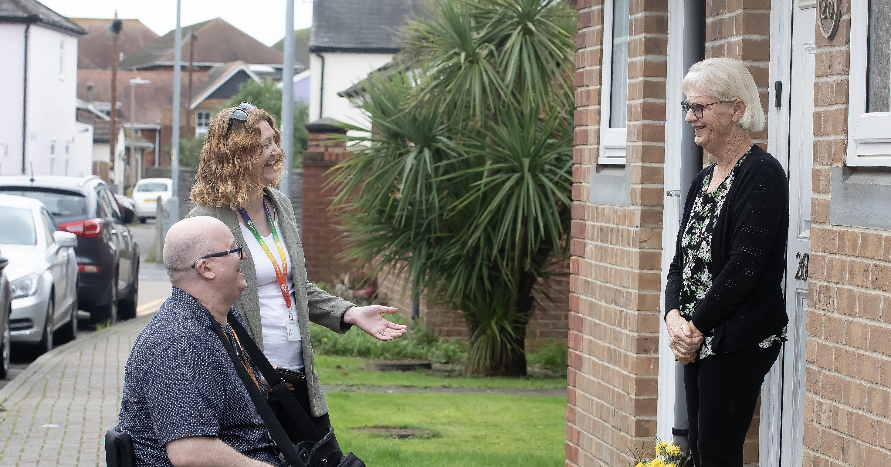 A woman is stood next to a man in a wheelchair. They are both talking to a woman who is stood on her doorstep.