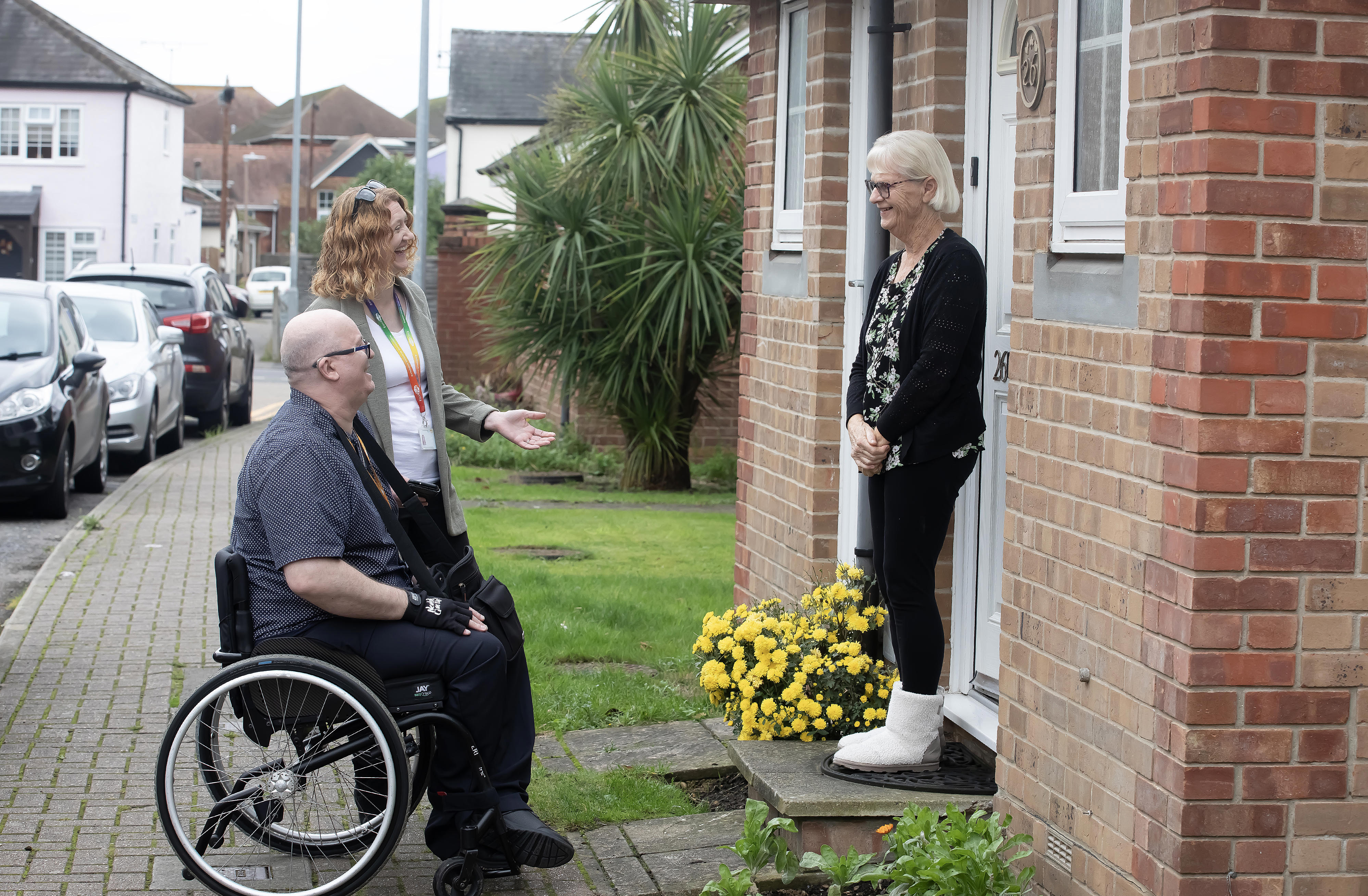 A woman is stood next to a man in a wheelchair. They are both talking to a woman who is stood on her doorstep.