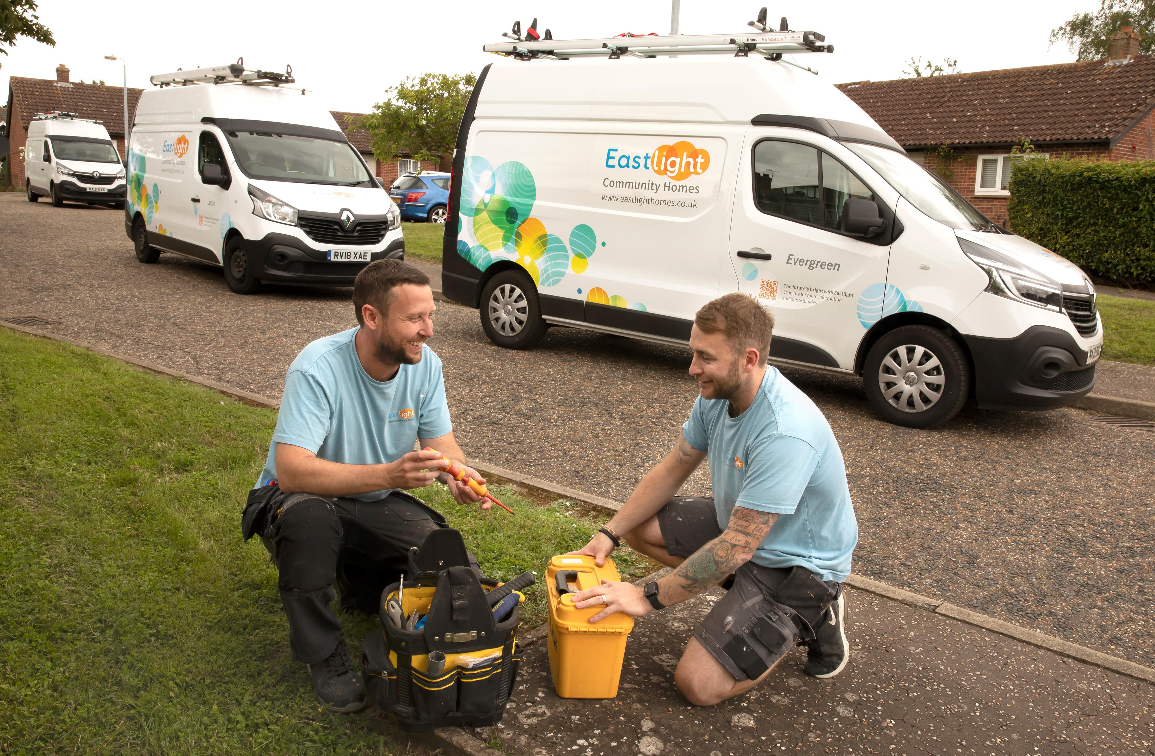 Two Eastlight tradesmen are kneeling at a toolbox on some grass. There are Eastlight vans parked on the road behind them.