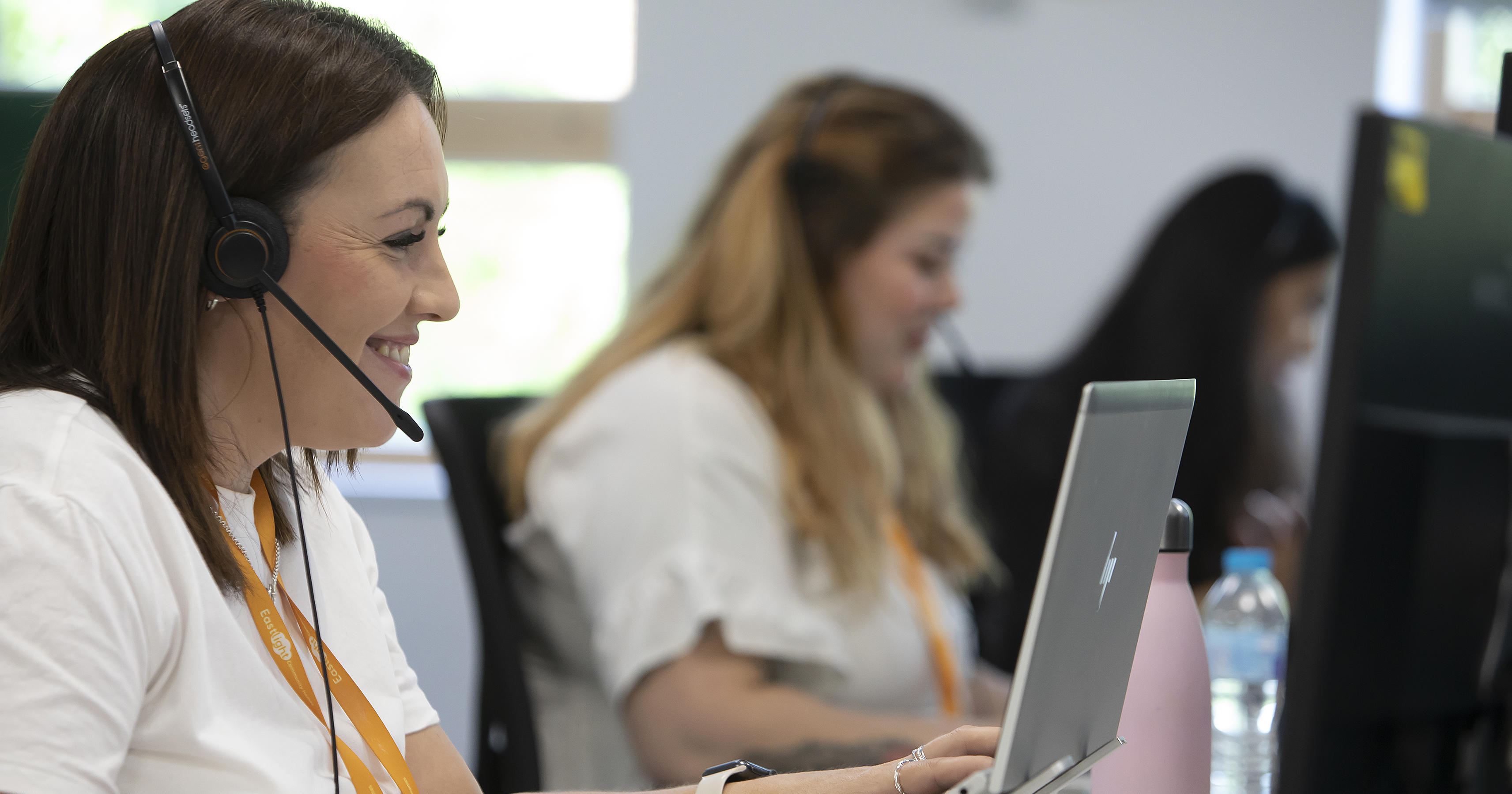 A woman wearing a headset is smiling at a laptop screen. There's another woman in the background doing the same.