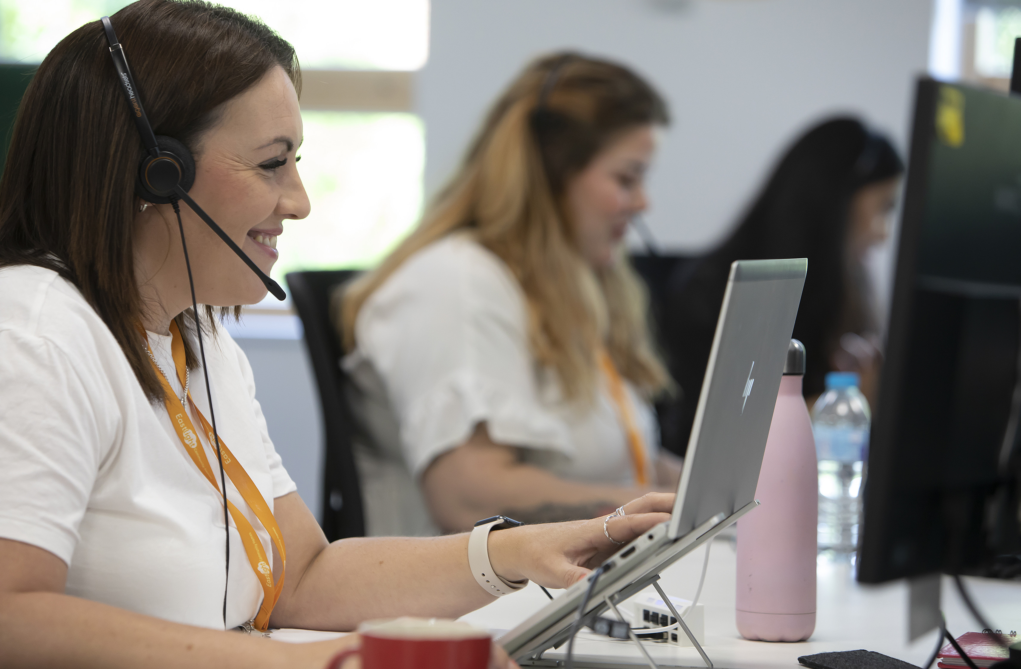 A woman wearing a headset is smiling at a laptop screen. There's another woman in the background doing the same.