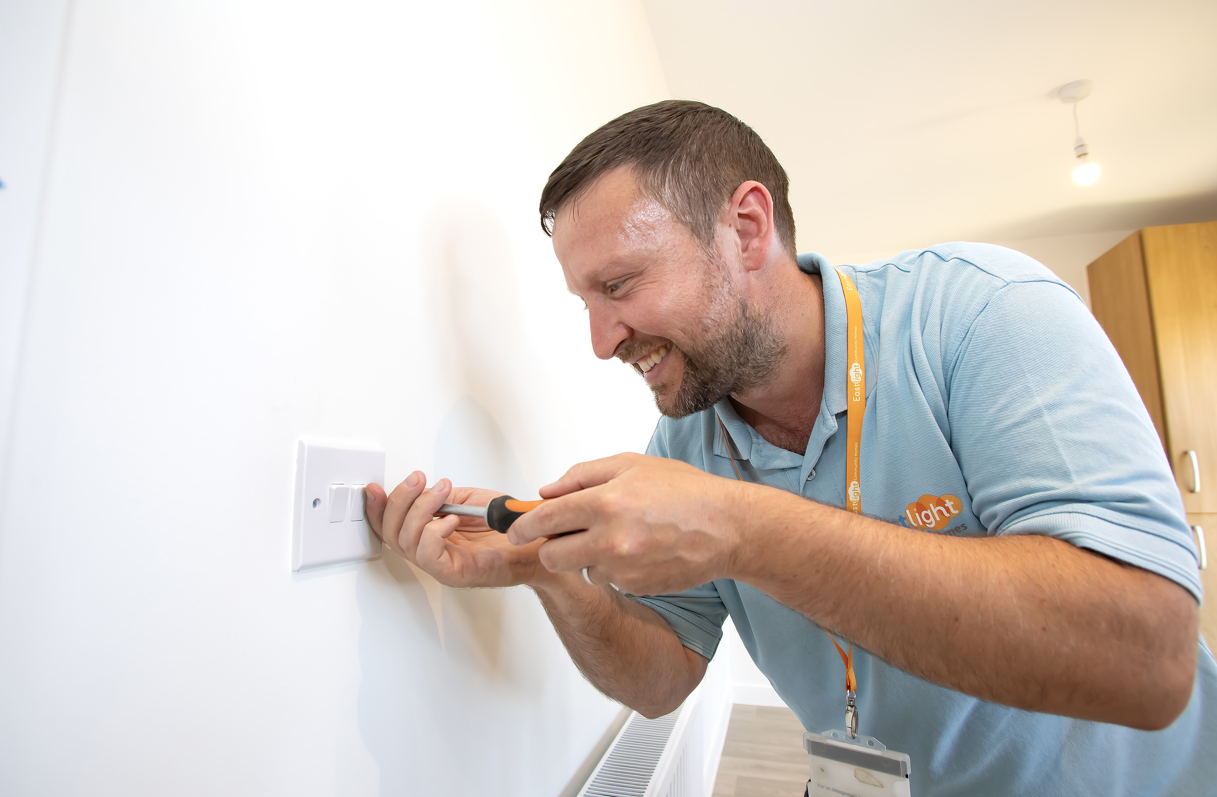 A man wearing a blue Eastlight tshirt is screwing a light switch to a wall.