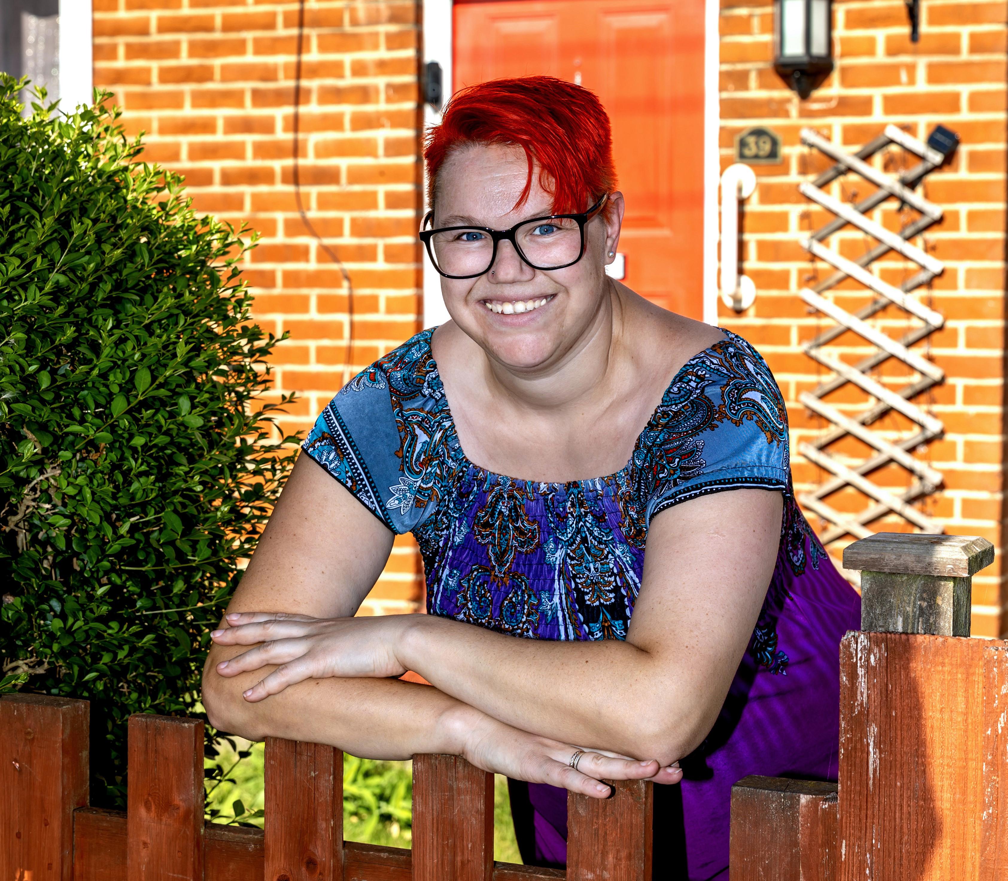 A woman with short red hair and glasses is leaning on a wooden gate in her front garden.