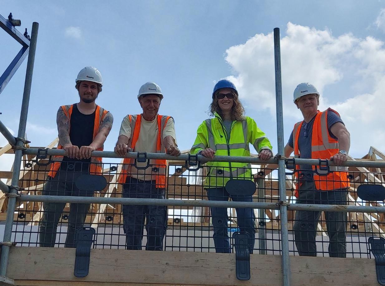 Eastlight Community Homes Development Manager with partners on a building site. Scaffolding is erected, and four people in hard hats and hi vis look down at the camera holding onto a railing and smiling