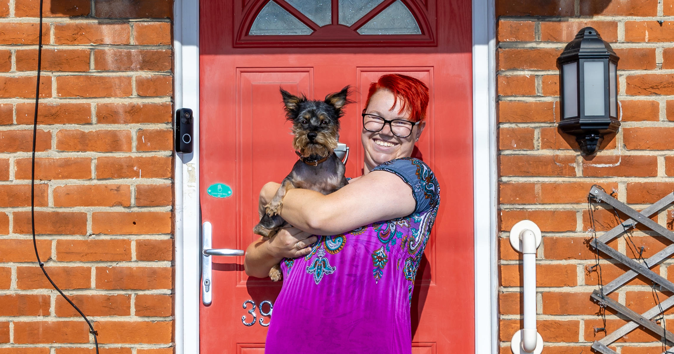 A woman is stood on her doorstep in front of a red front door. She is holding a small yorkshire terrier in her arms.
