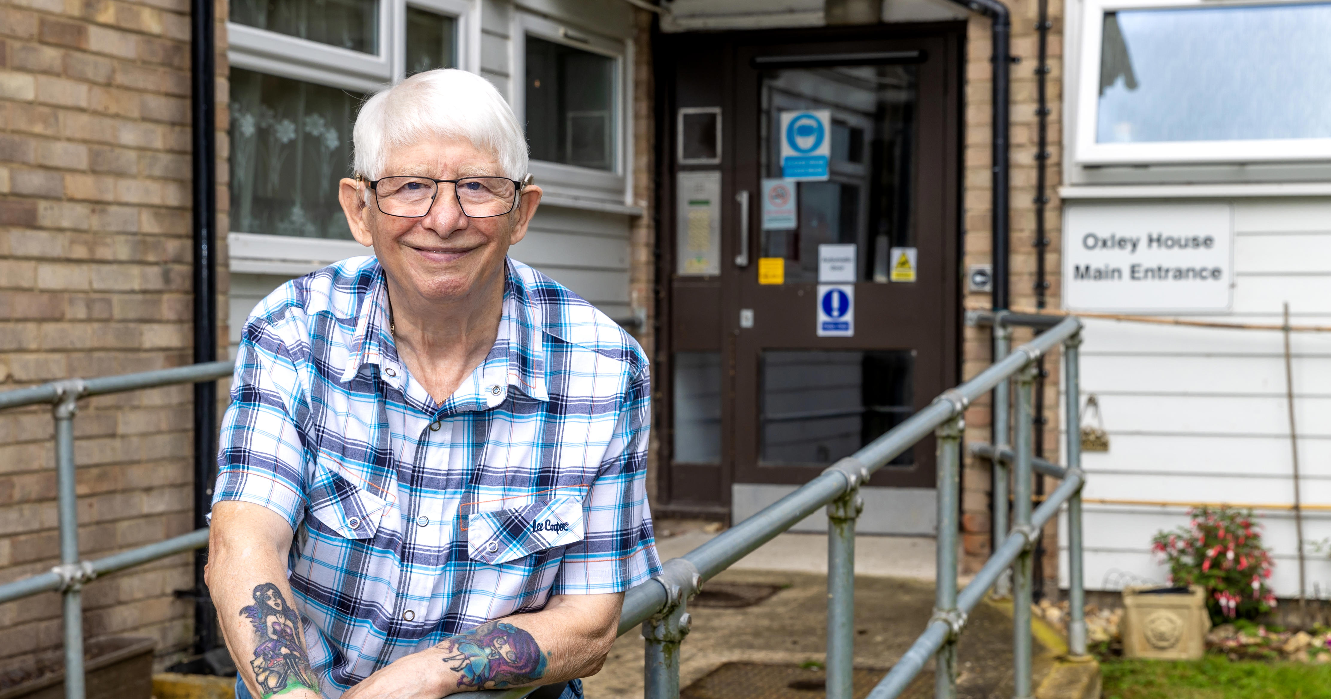 An older man with white hair and glasses is leaning on a metal railing which leads up to a block of flats.
