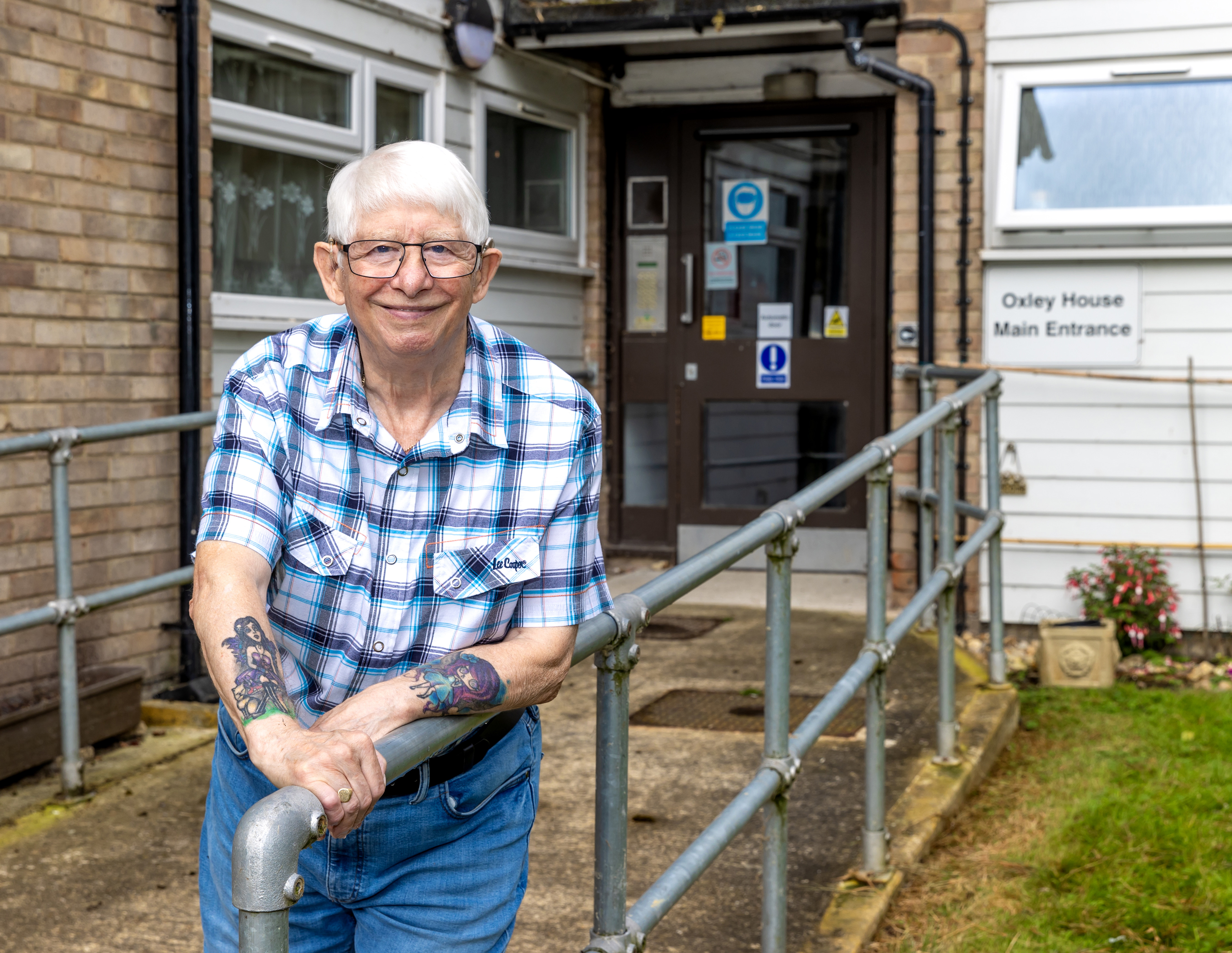 An older man with white hair and glasses is leaning on a metal railing which leads up to a block of flats.