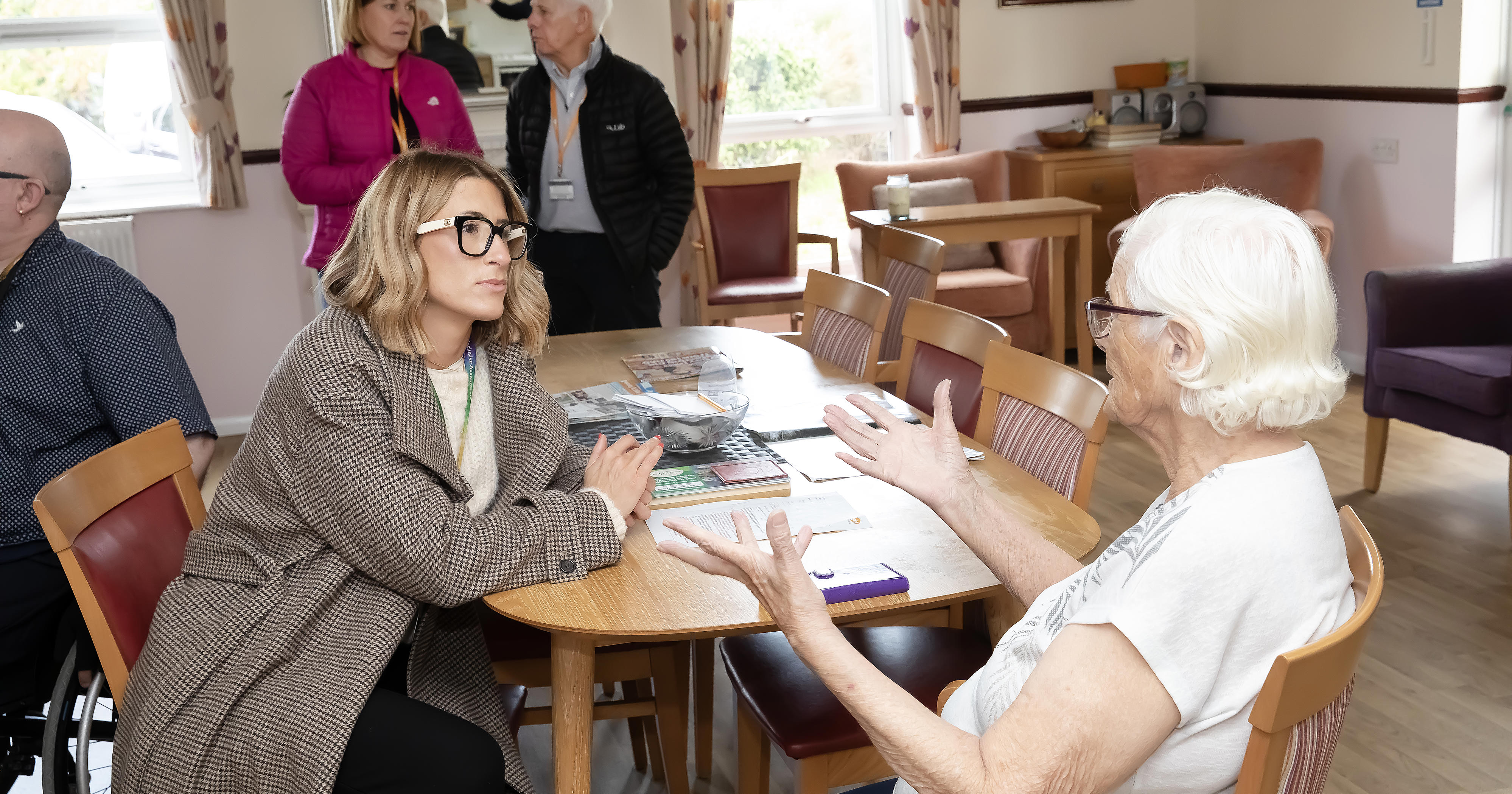 Two women, one with white hair, are sat at a table talking. A man and woman are in the background also talking.