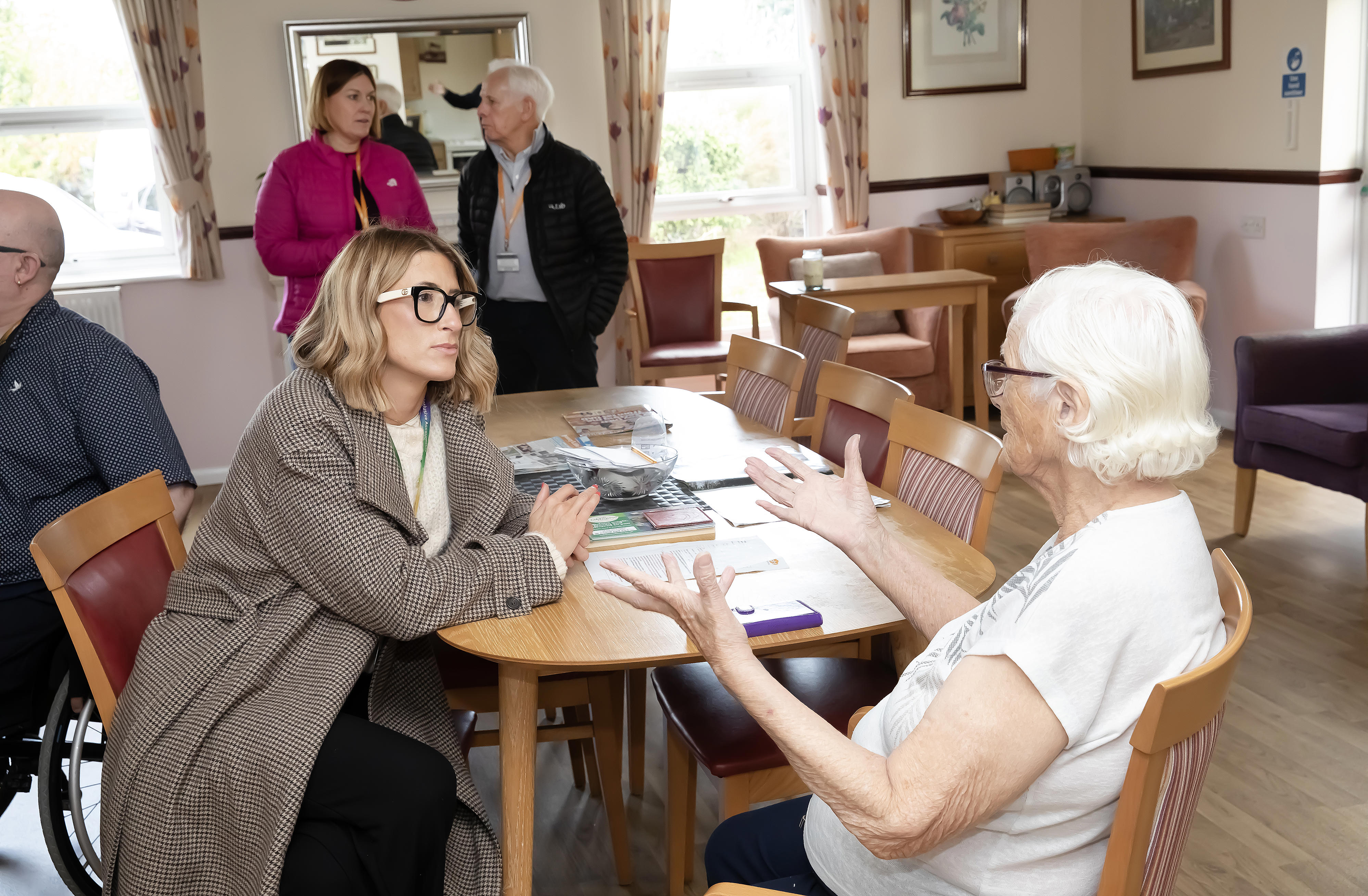 Two women, one with white hair, are sat at a table talking. A man and woman are in the background also talking.