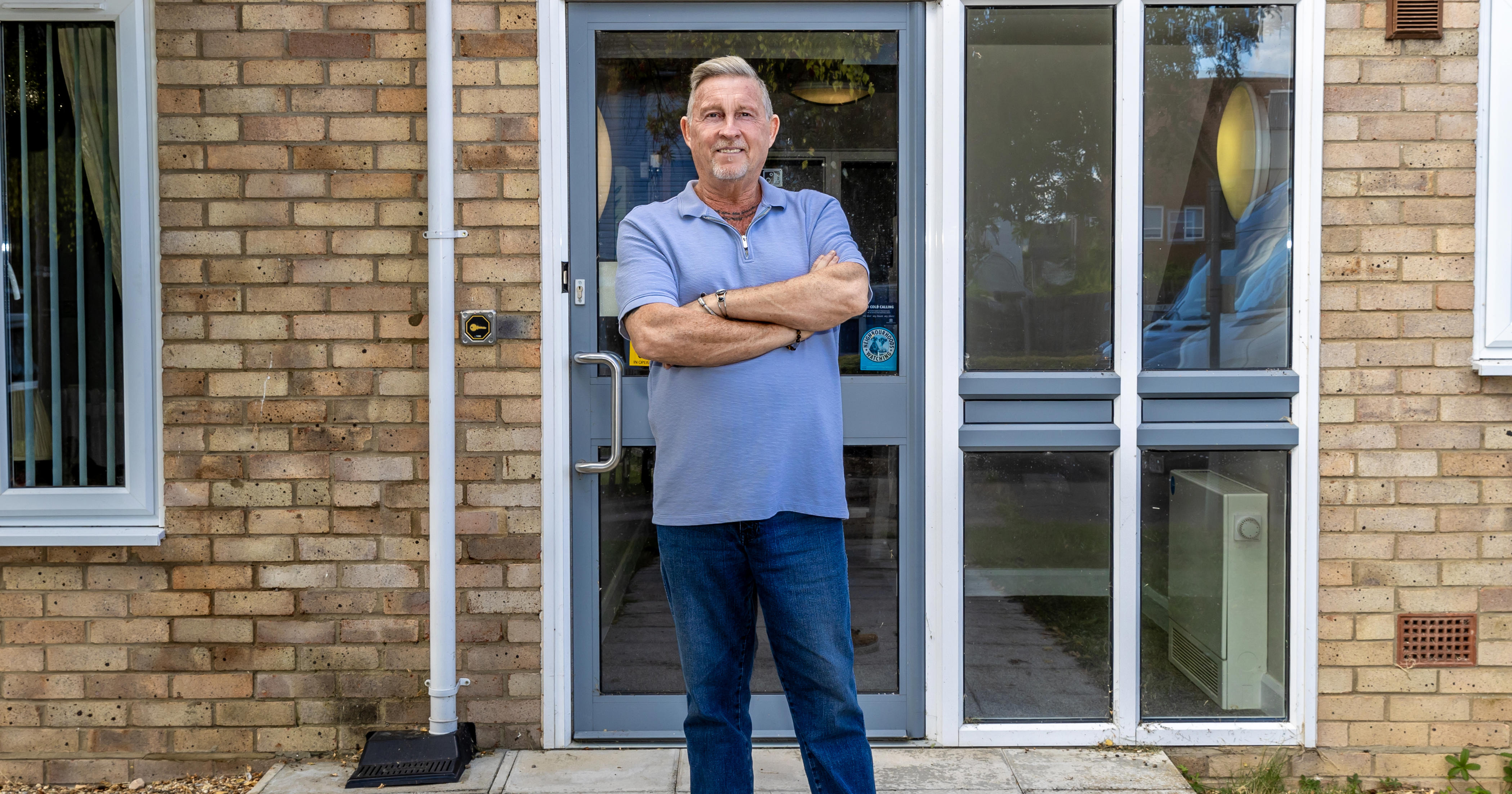 A man is stood with his arms folded in front of a door to flats.