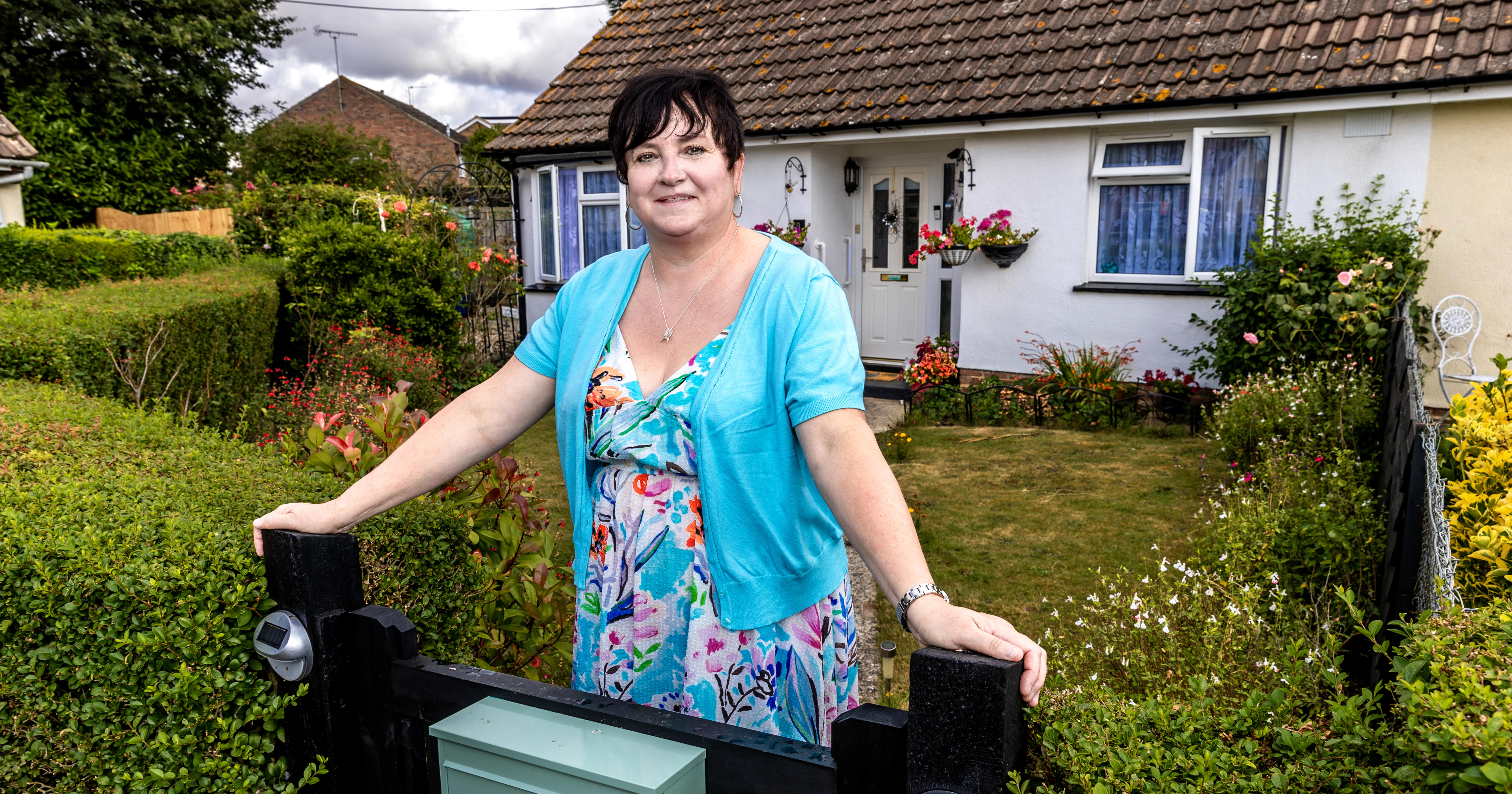 A woman in a blue floral dress is holding onto two gate posts to the entry of her garden gate. She is smiling at the camera.