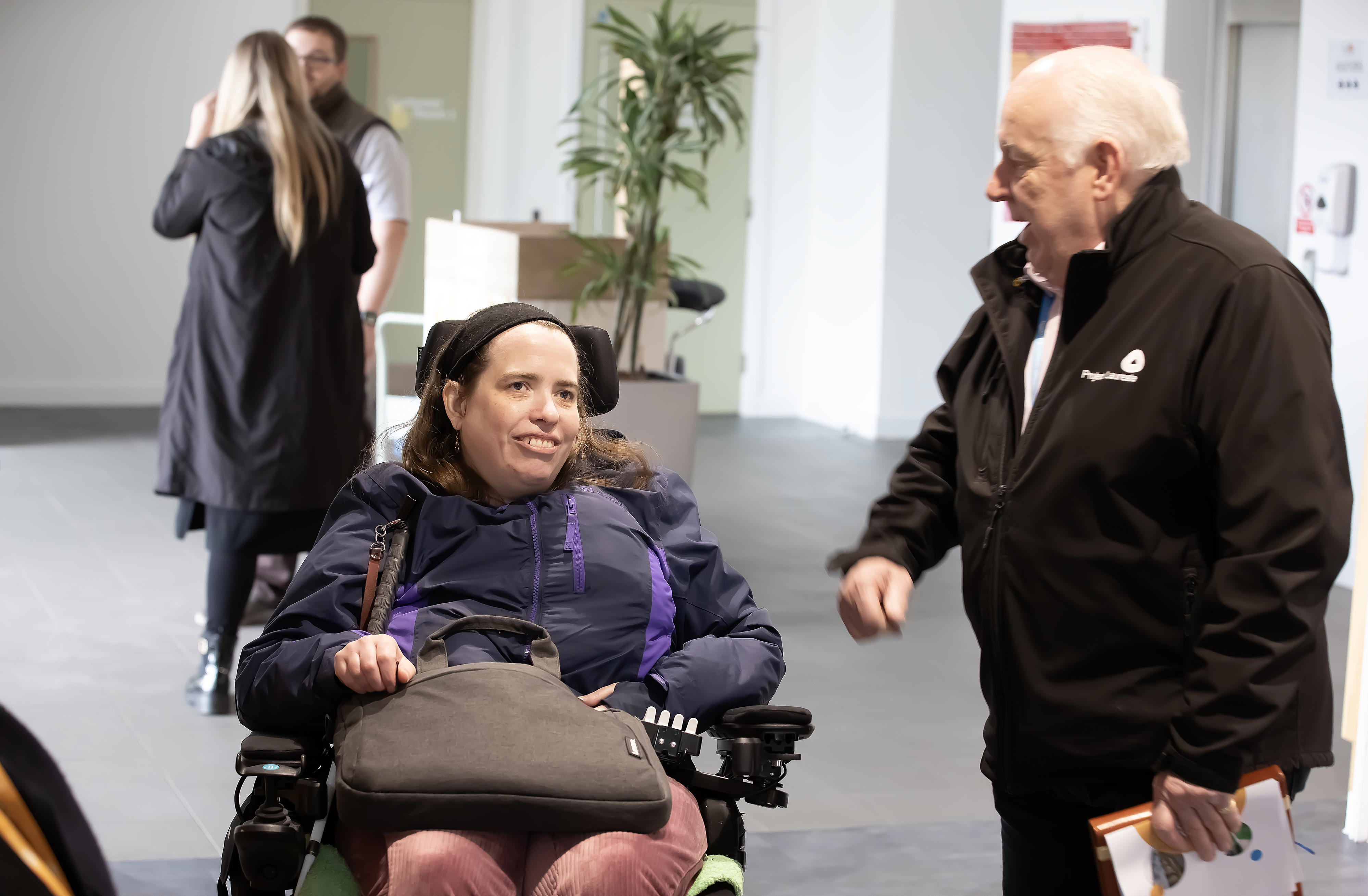 A woman in a wheelchair is speaking to a man stood next to her.