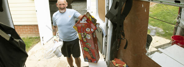 A man holding cardboard boxes and a fan. He is standing at the back of a van putting the items into the van.