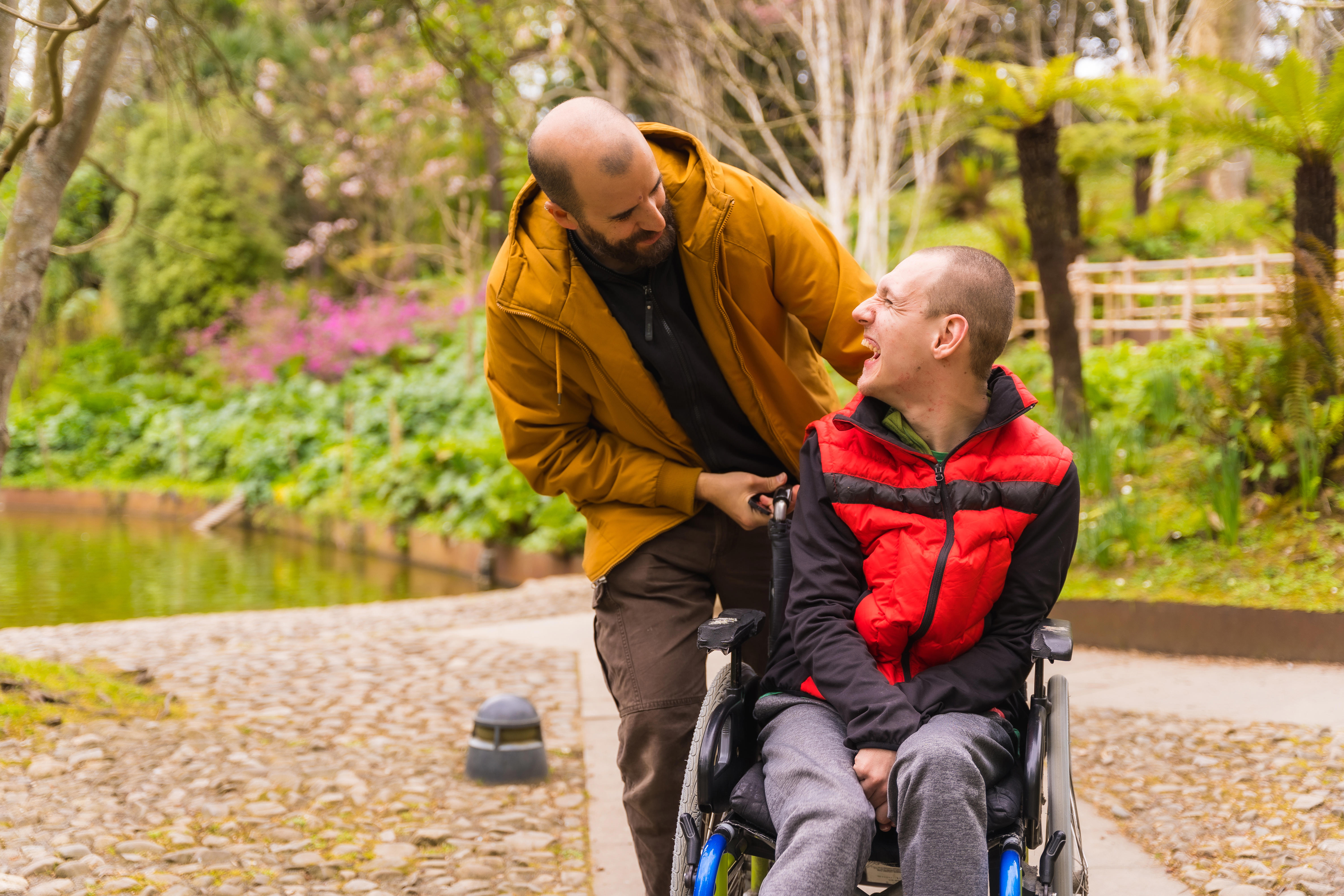 A man in a wheelchair is being pushed by a male carer. They are both laughing.