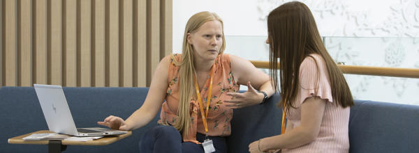 Two women are sat on a sofa together. One woman is talking to the other, whilst the other looks at a laptop screen.