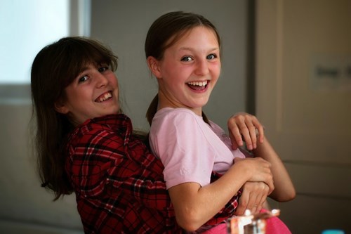 Two young girls smiling at the camera, one behind the other with arms around the other's body