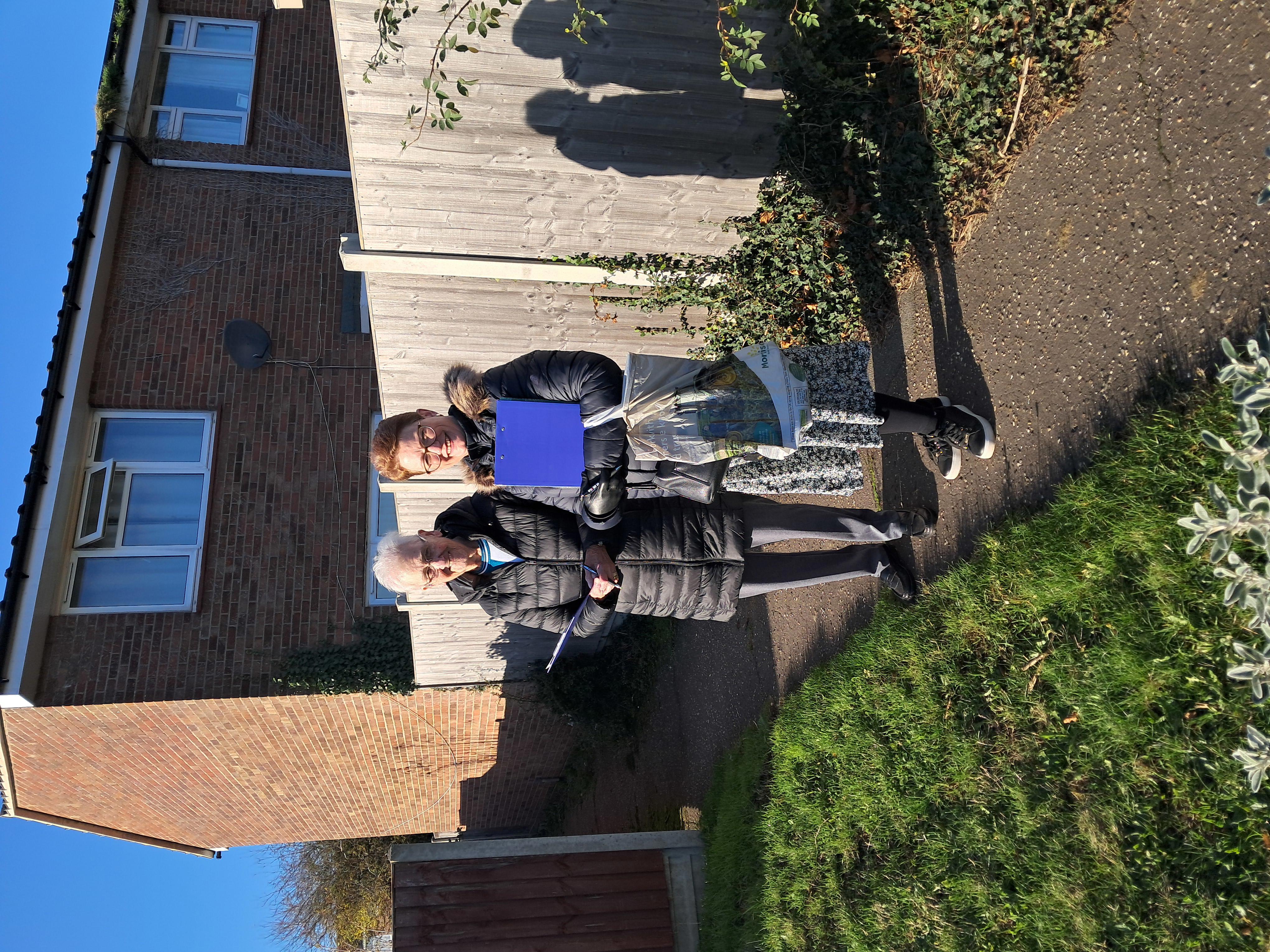 Two women are stood next to each other on a path beside a fence. They are both holding clipboards.