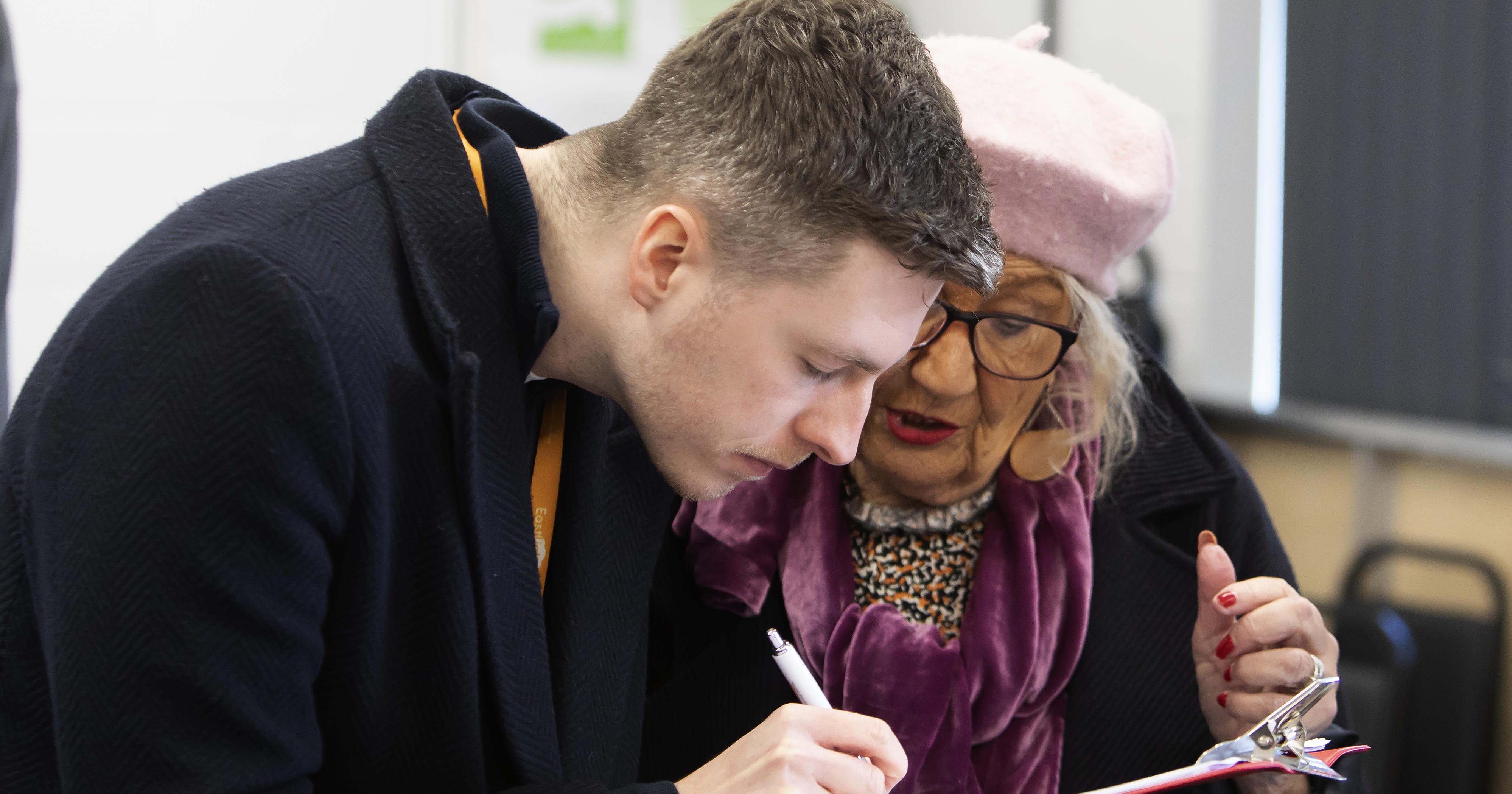 A man and a woman are stood together looking at a clipboard. The man is writing on the clipboard with a pen.