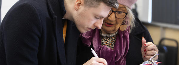 A man and a woman are stood together looking at a clipboard. The man is writing on the clipboard with a pen.