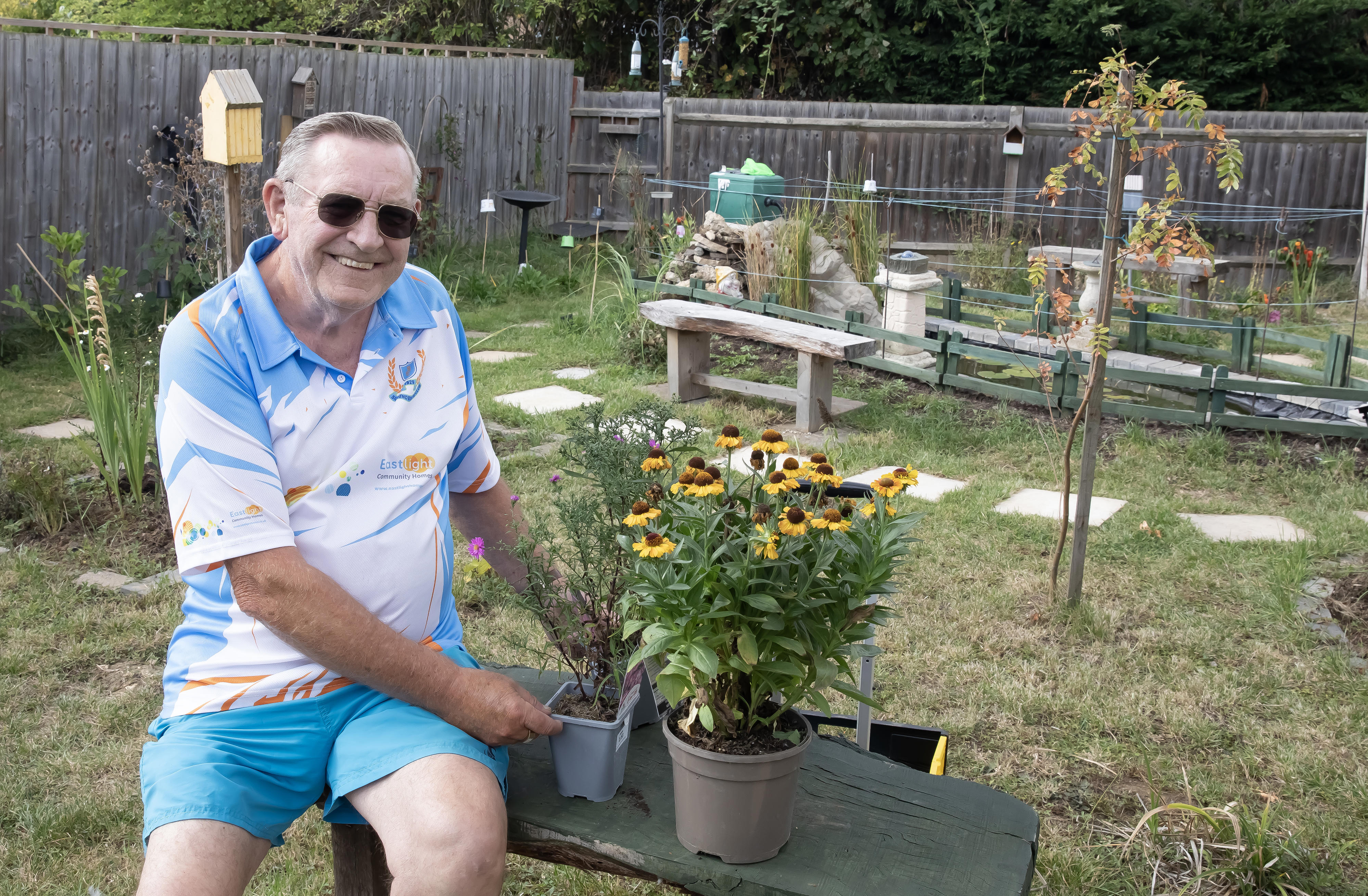 A man is sitting on a bench in a garden. On the bench beside him is a pot of yellow flowers.