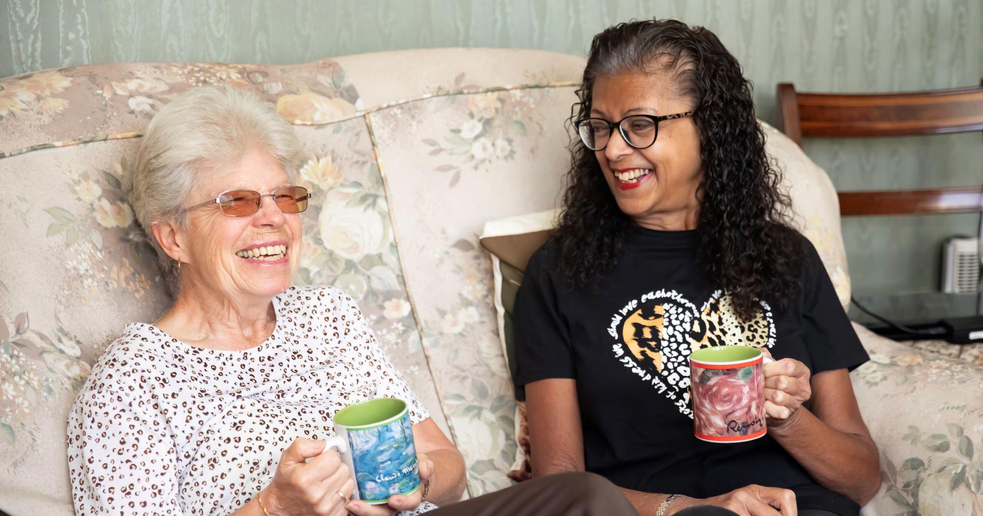 Two women sat on a sofa holding mugs of tea. They are both laughing and smiling.