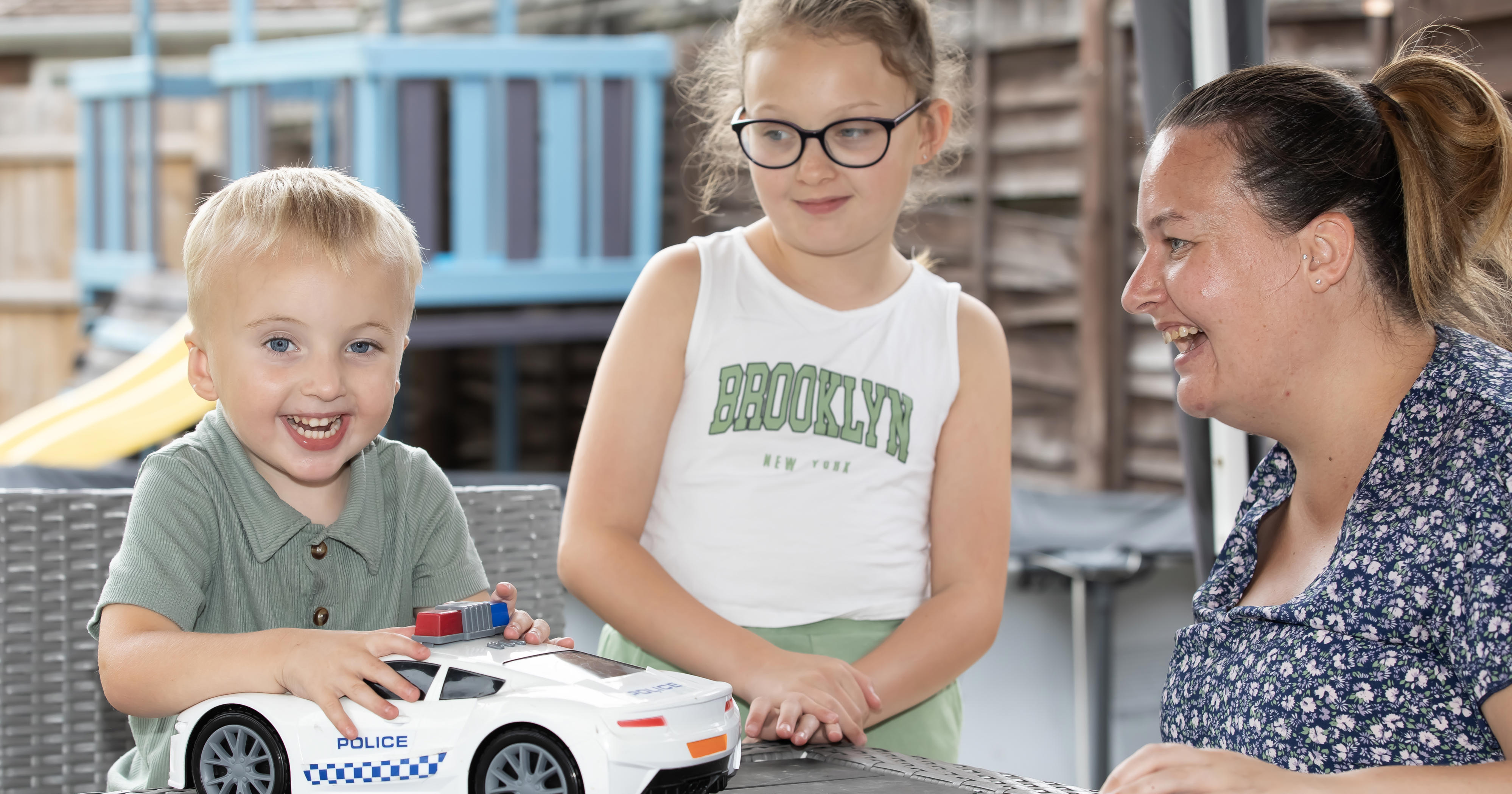 A woman with her two young children are playing on a table. The young boy is holding a large police car toy.