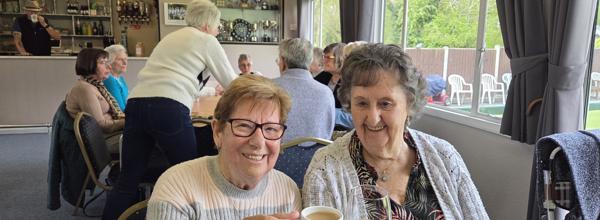 Two older ladies are sat beside each other cheersing their cups of tea together.